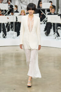 Model walks the runway in a white lace-front jumpsuit, with an orchestra of musicians in black attire in the background.