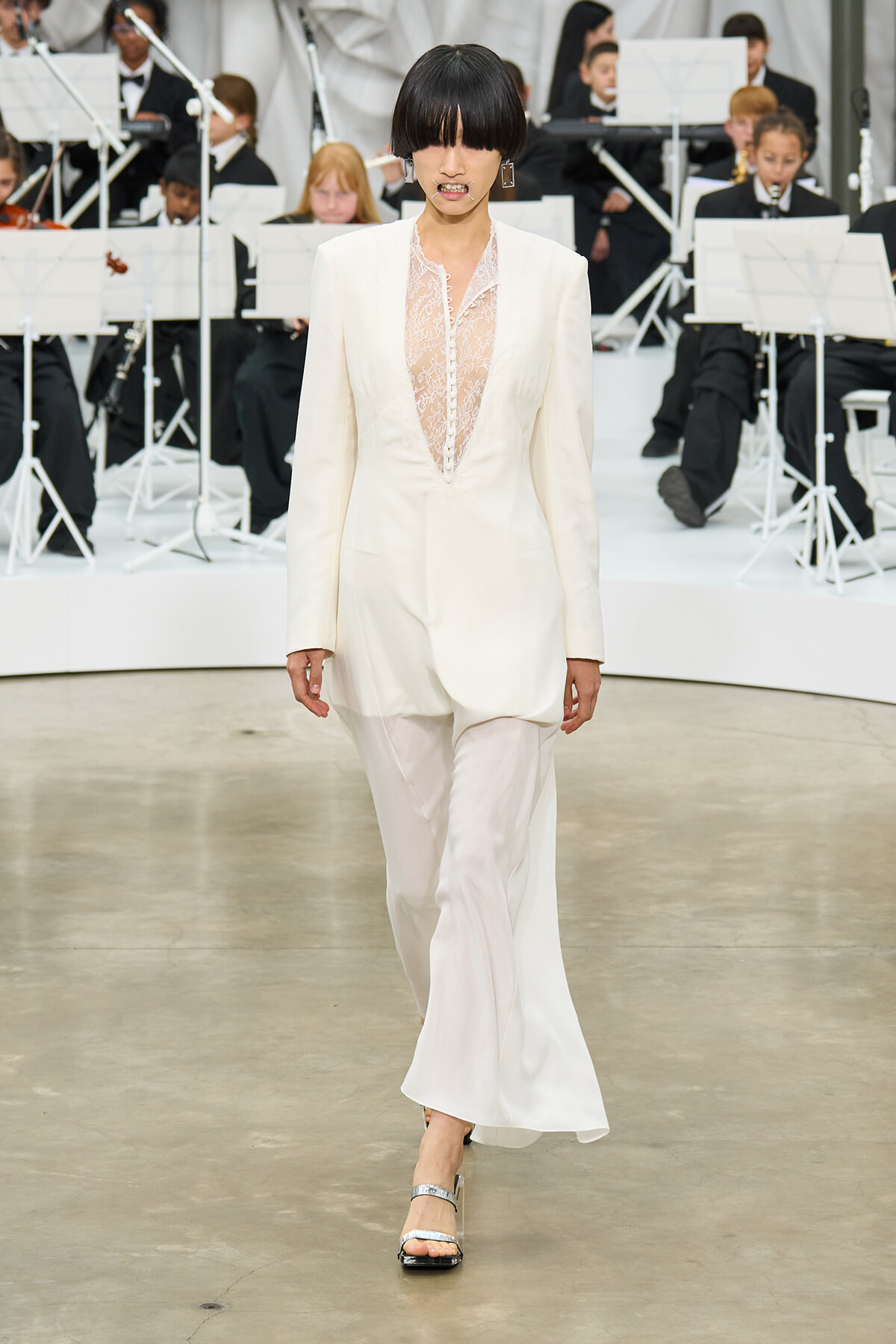 Model walks the runway in a white lace-front jumpsuit, with an orchestra of musicians in black attire in the background.