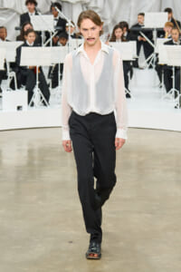 Model walking a runway in a sheer white blouse and black dress pants, with an orchestra in the background.