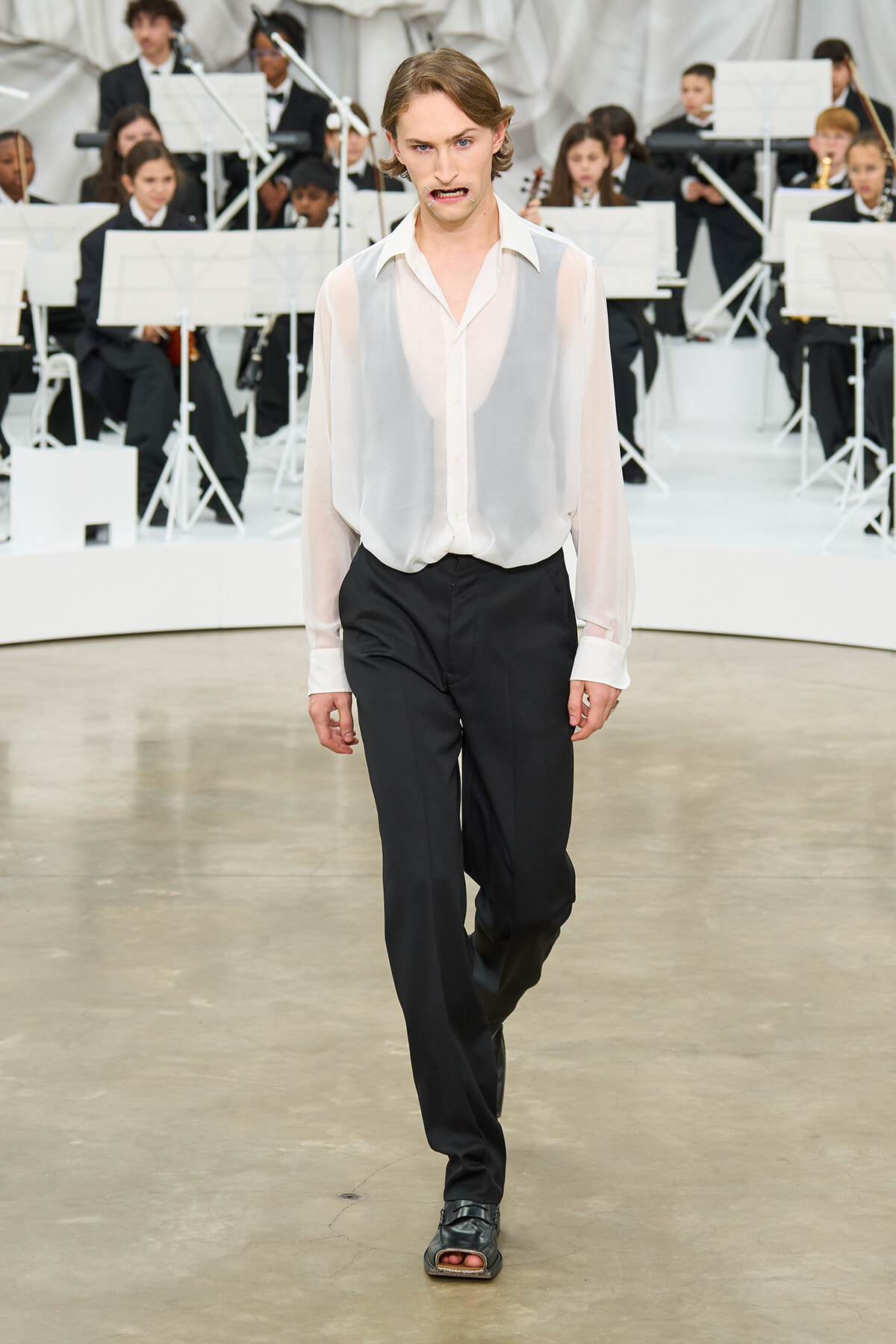 Model walking a runway in a sheer white blouse and black dress pants, with an orchestra in the background.