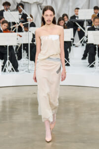 Model walking the runway in a pale beige slip dress with lace trim, orchestra in background.