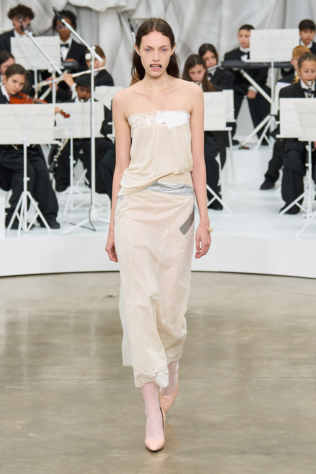 Model walking the runway in a pale beige slip dress with lace trim, orchestra in background.