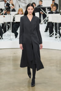 Female model walks a runway in a long charcoal gray tailored coat, with an orchestra visible in the background behind her?no music notes? (stage scene).