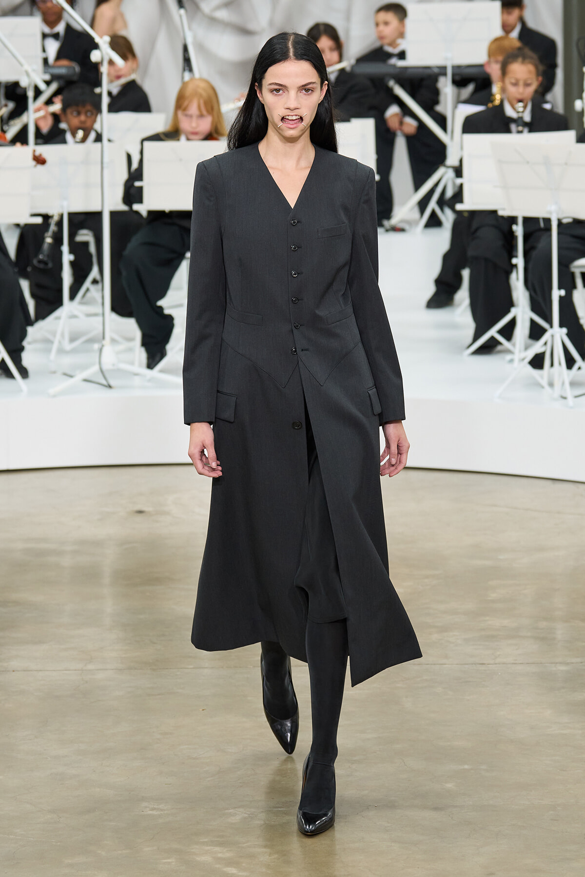 Female model walks a runway in a long charcoal gray tailored coat, with an orchestra visible in the background behind her?no music notes? (stage scene).