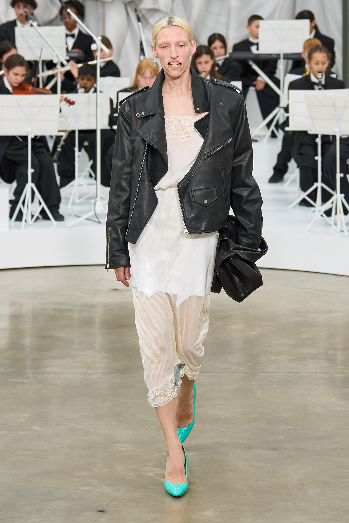 Model walking a runway in a black leather biker jacket over a cream lace-top dress and sheer pants, teal heels, orchestral musicians in background.