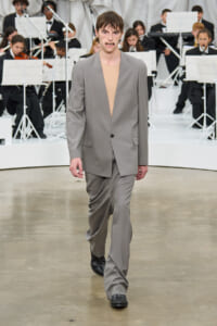 Male model walks a runway in a loose gray suit with a beige top, orchestra visible in the background.