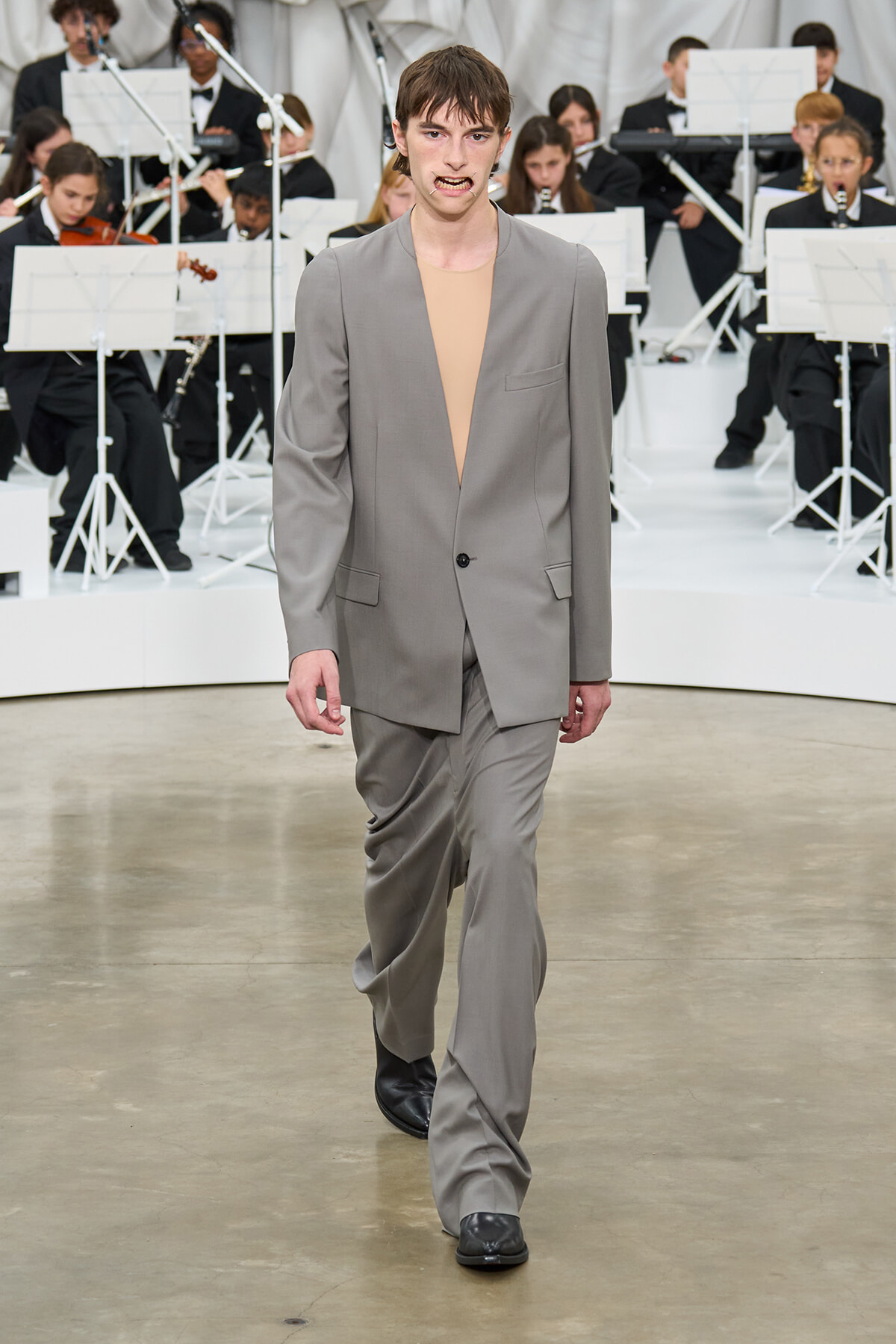 Male model walks a runway in a loose gray suit with a beige top, orchestra visible in the background.
