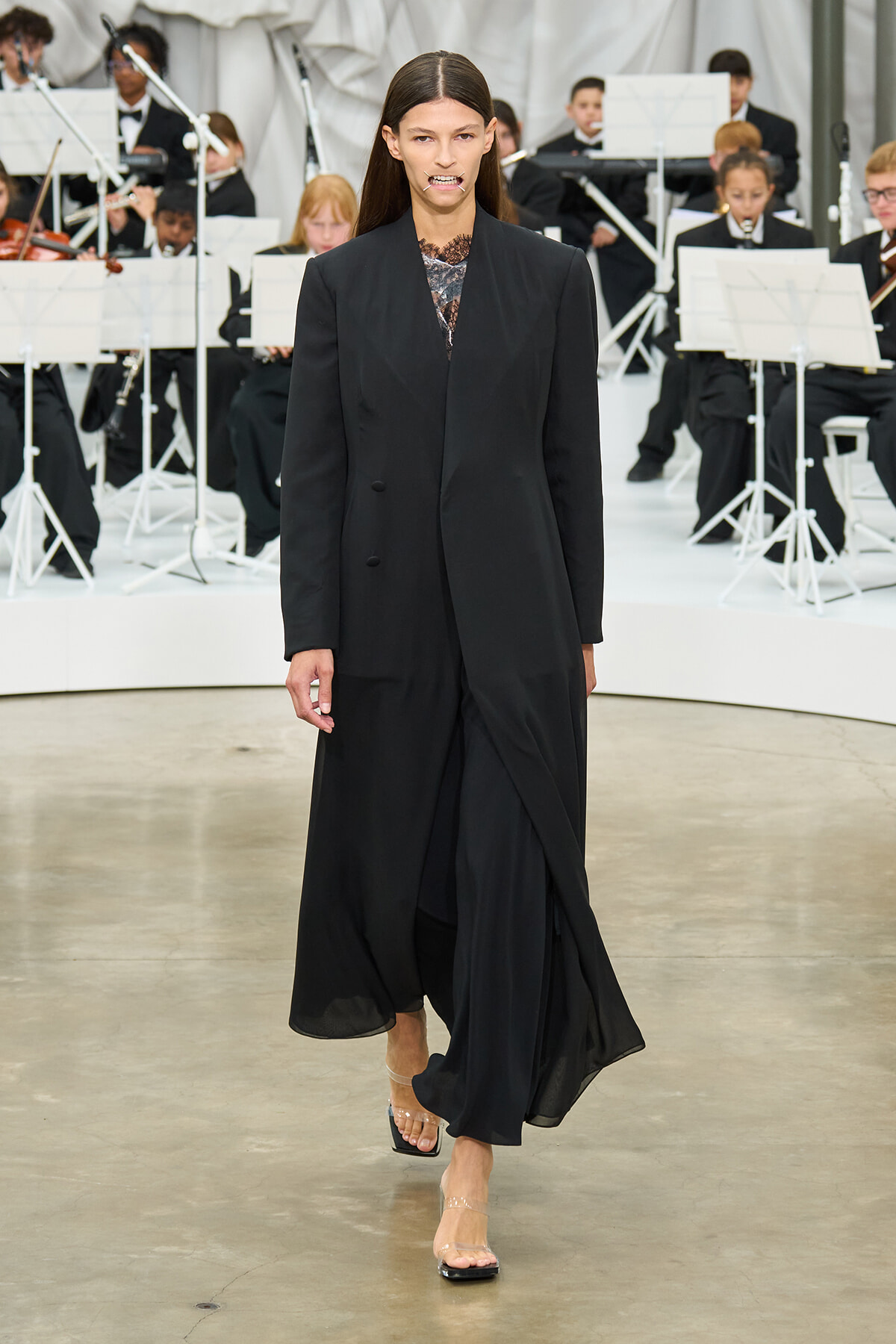 Model walking a fashion show in a black long coat over a lace top; orchestra musicians in the background.