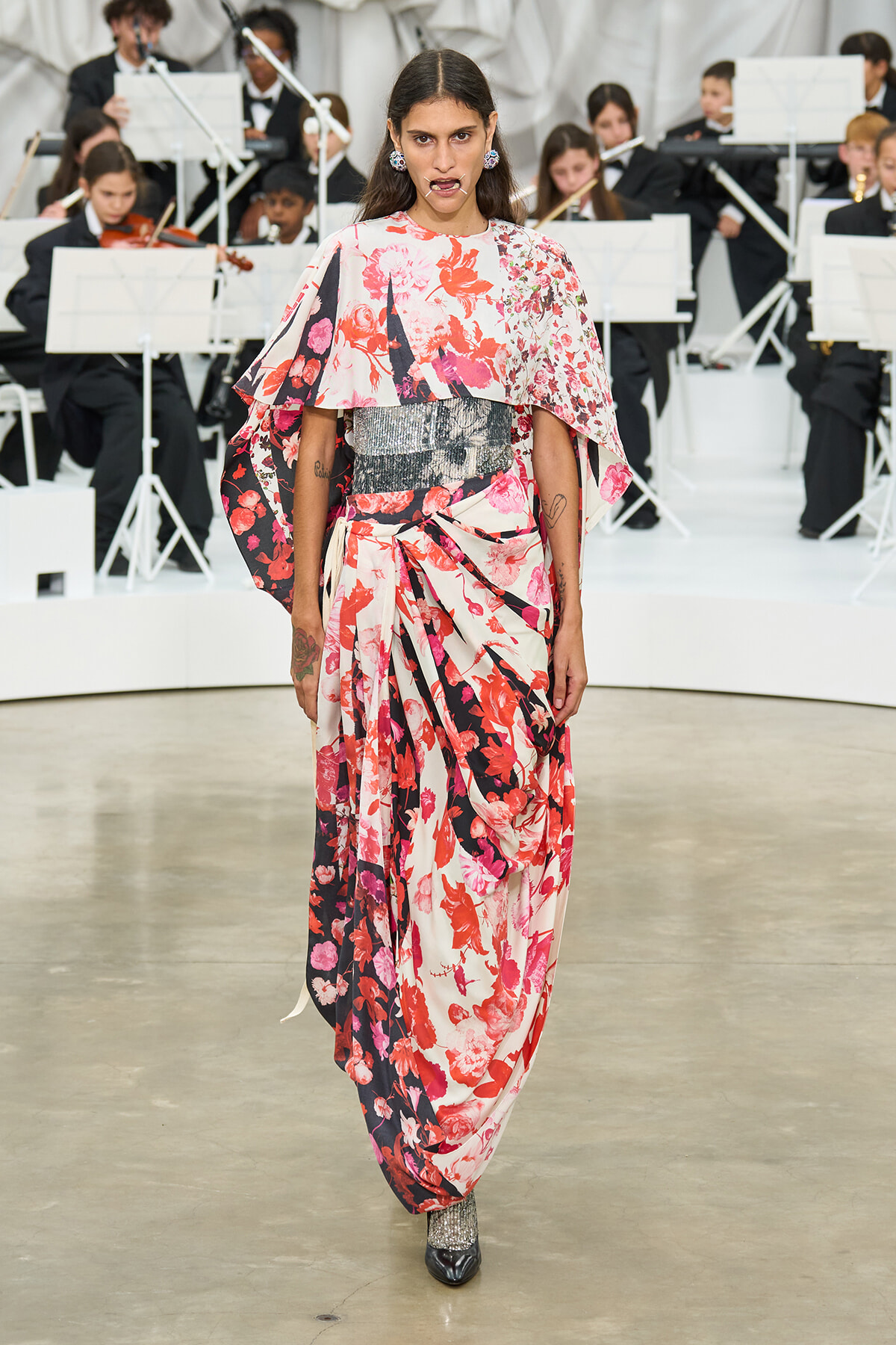 Model walking a runway in a pink-red floral cape dress with a metallic belt, an orchestra visible in the background