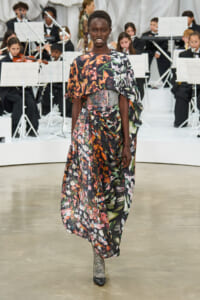 Model walks a floral maxi dress with a jeweled waist belt on a runway, with an orchestra visible in the background.