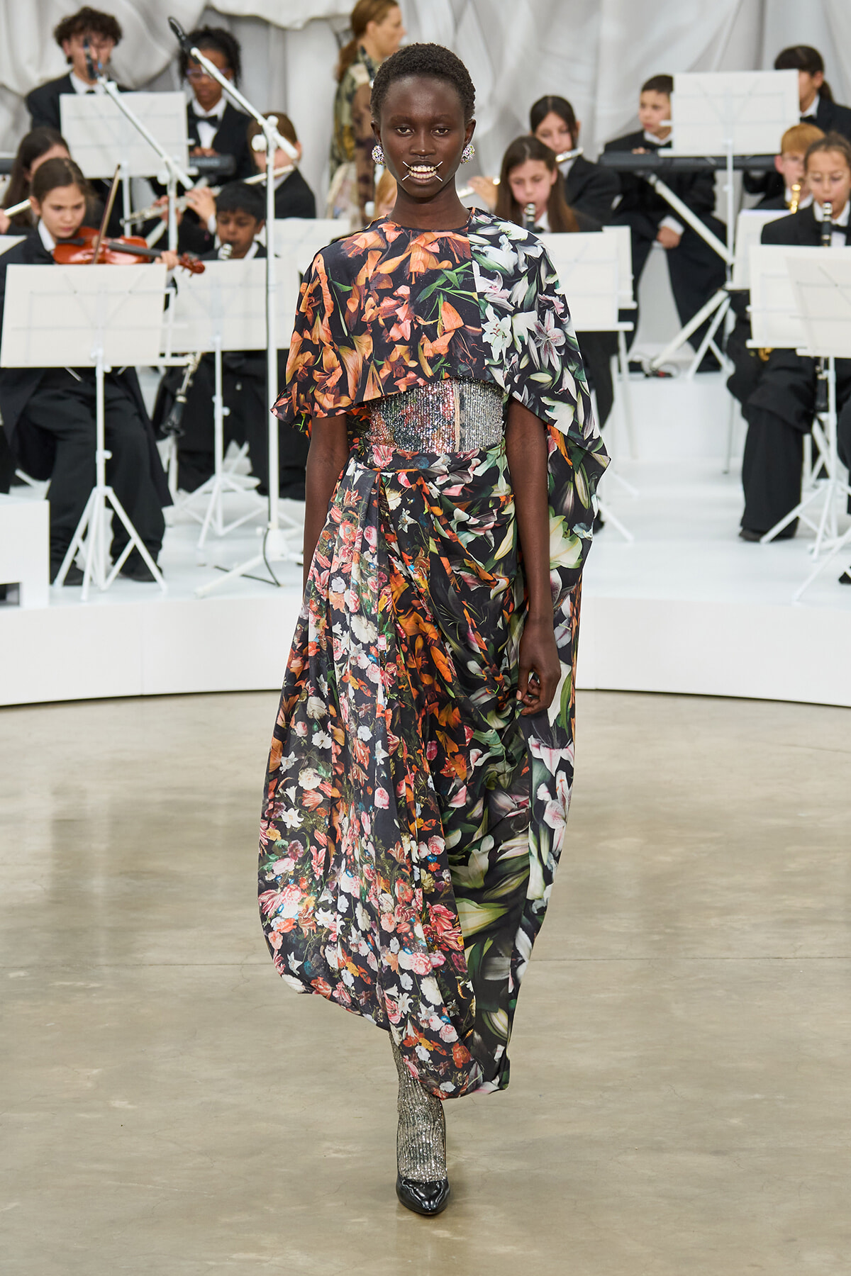 Model walks a floral maxi dress with a jeweled waist belt on a runway, with an orchestra visible in the background.