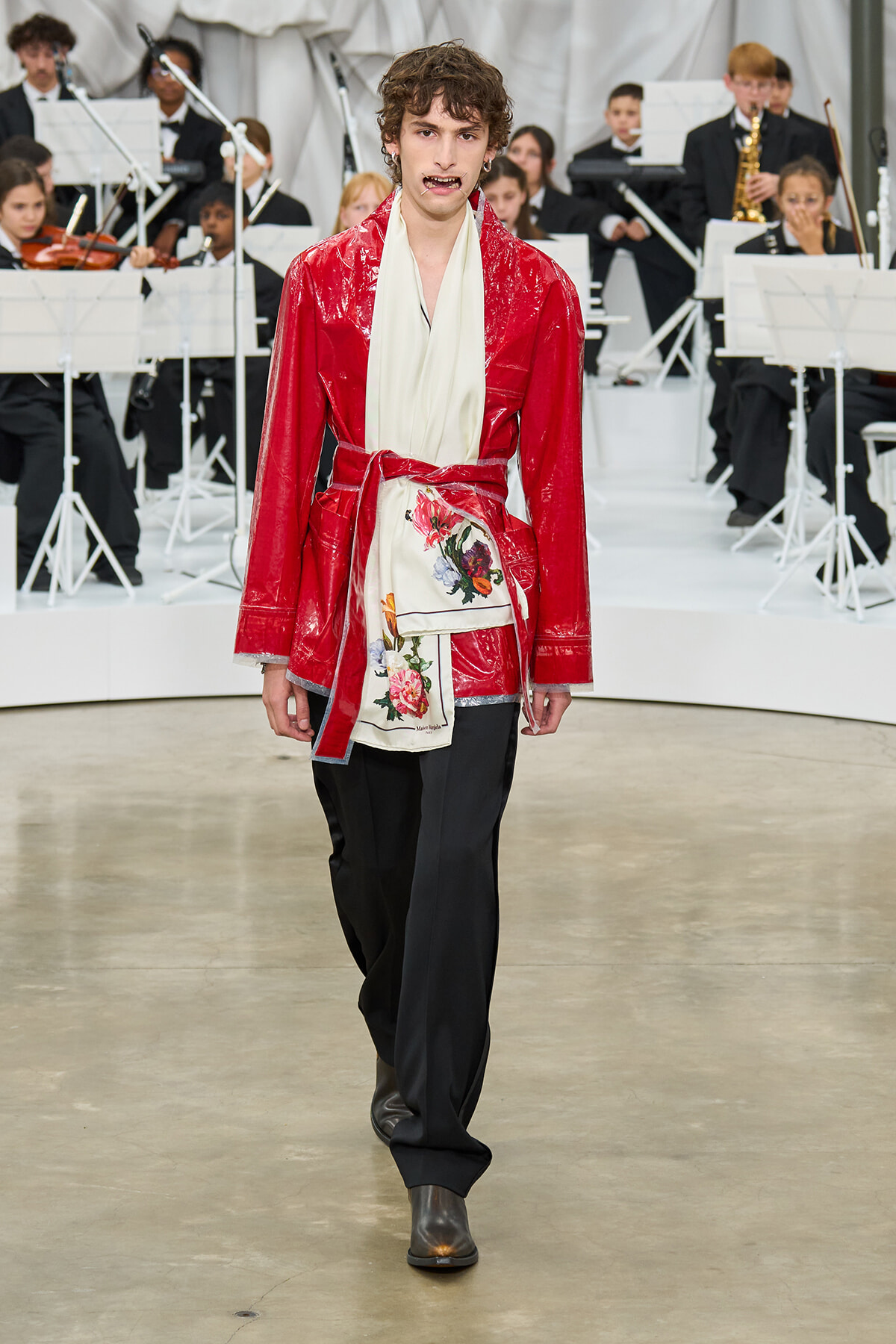 Male model walks a runway in a red glossy robe with a cream sash and floral-printed panel, during a fashion show with an orchestra in the background.