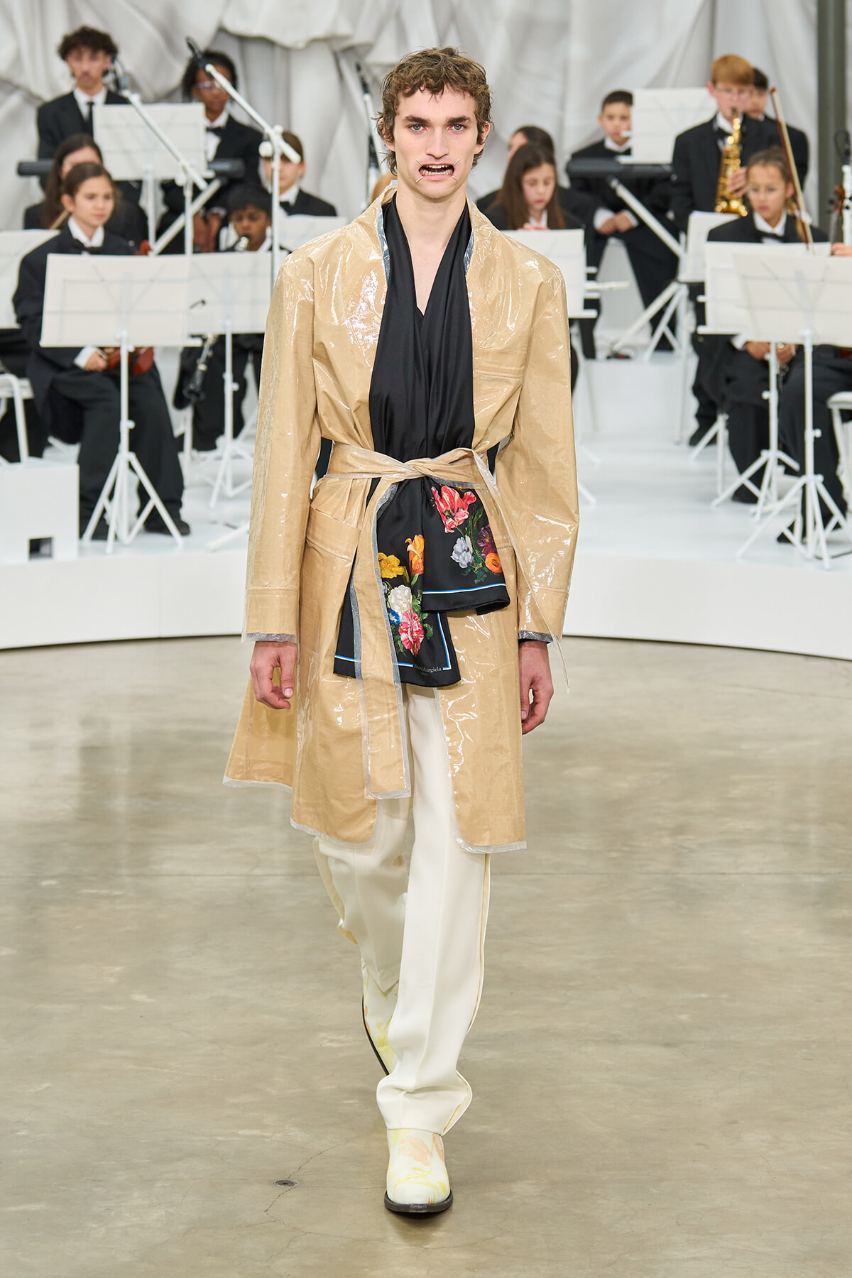 Male model walks the runway in a shiny beige trench with a black draped top and floral sash, white pants, and pale patterned shoes, with an orchestra in the background.