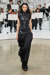 Model walks the runway in a shiny black sleeveless top and draped black skirt; an orchestra performs in the background.