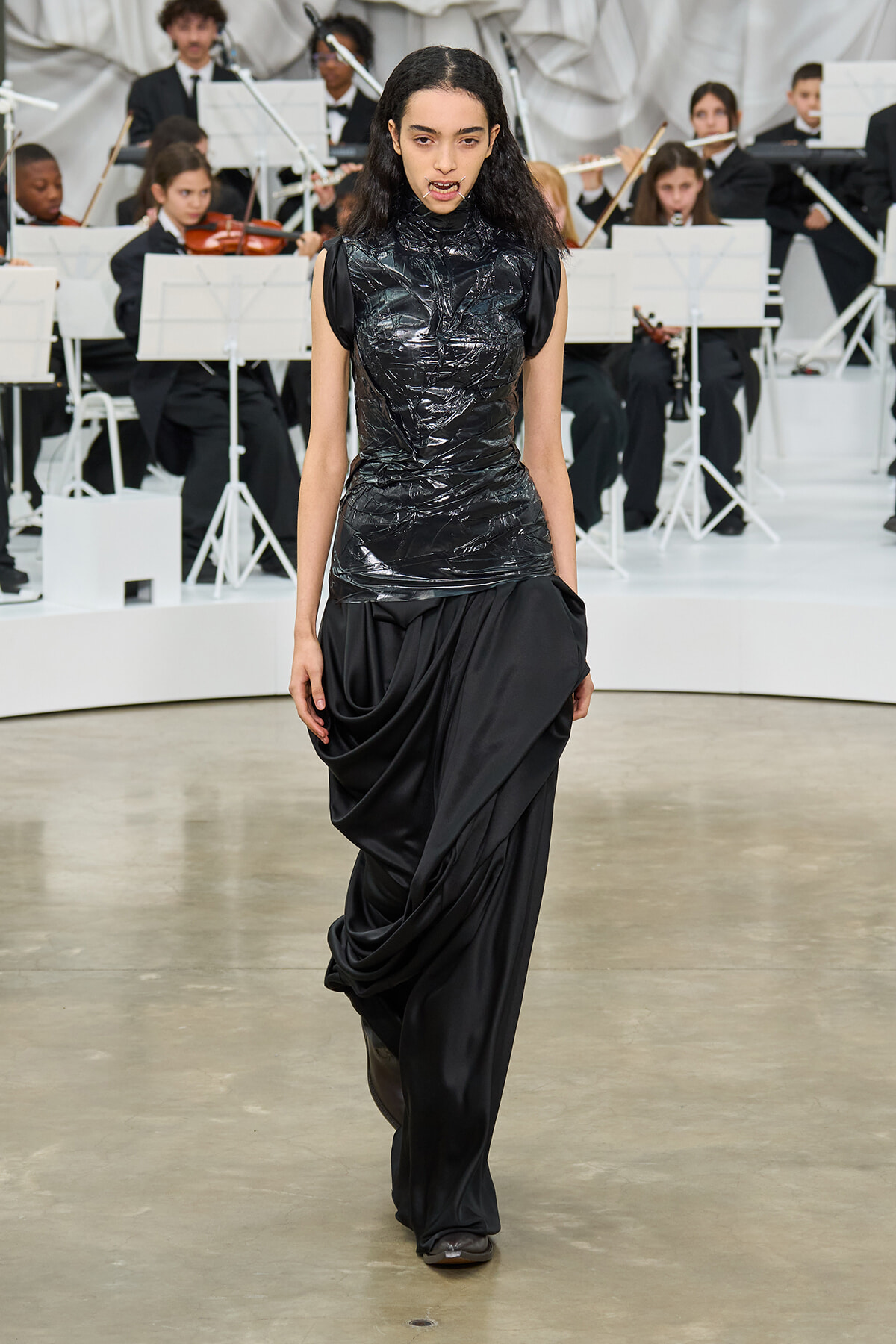 Model walks the runway in a shiny black sleeveless top and draped black skirt; an orchestra performs in the background.