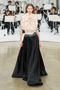 Model walking the runway in a cream ruffled blouse and long black satin skirt, with an orchestra in the background.