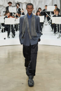 Model walking a fashion runway in a blue-gray pinstripe blazer, matching scarf, oversized black pants, and chunky strapped shoes; a formal orchestra audience in the background.