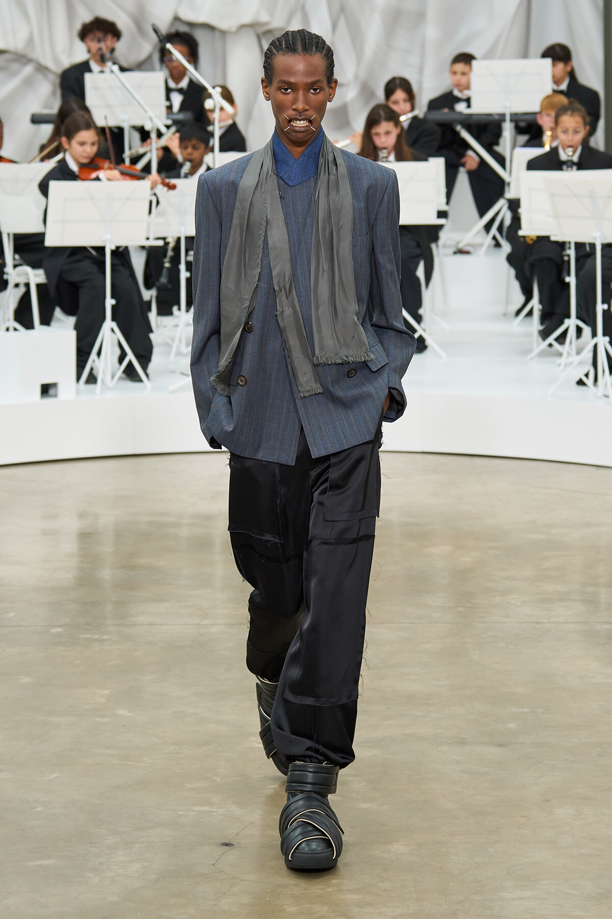 Model walking a fashion runway in a blue-gray pinstripe blazer, matching scarf, oversized black pants, and chunky strapped shoes; a formal orchestra audience in the background.