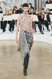 Model walking the runway in a crystal-covered sheer bodice and a metallic, draped skirt; an orchestra sits in the background of a fashion show.