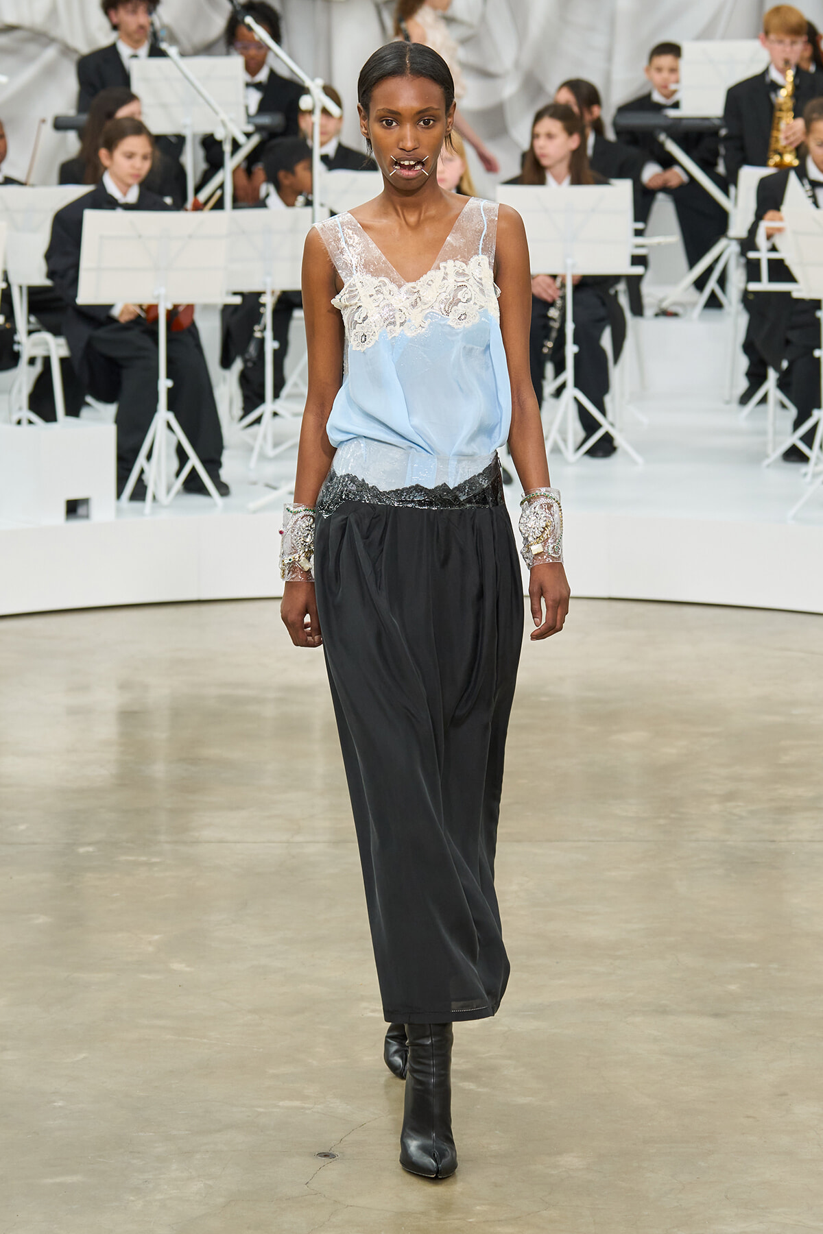 Model walking a runway in a light blue lace-trim top and black wide-leg pants, with silver embellished cuffs, as a live orchestra plays in the background.
