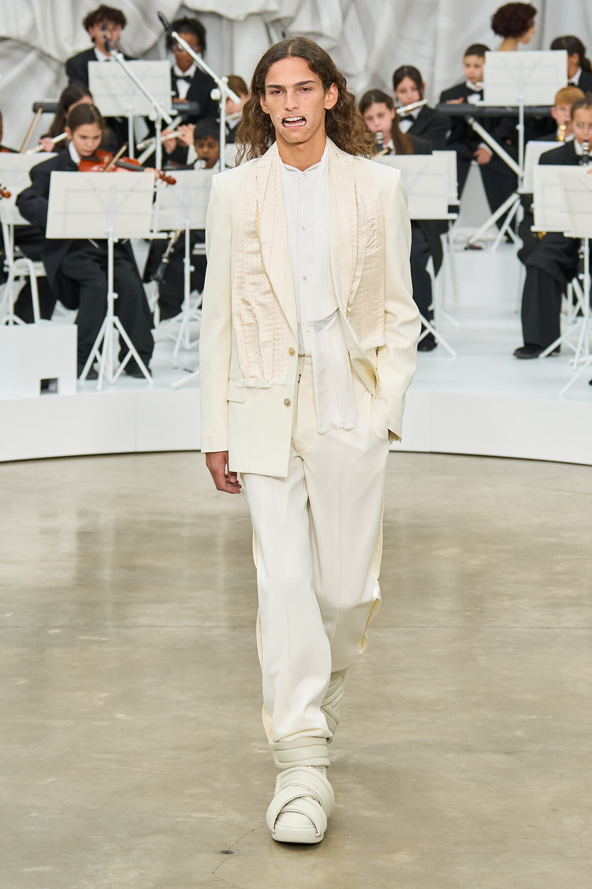 Model in an ivory suit with a scarf walks the runway as a white-clad orchestra performs behind.