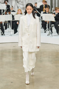 Model in a glossy white suit and matching pants walks a runway, with an orchestra visible in the background.