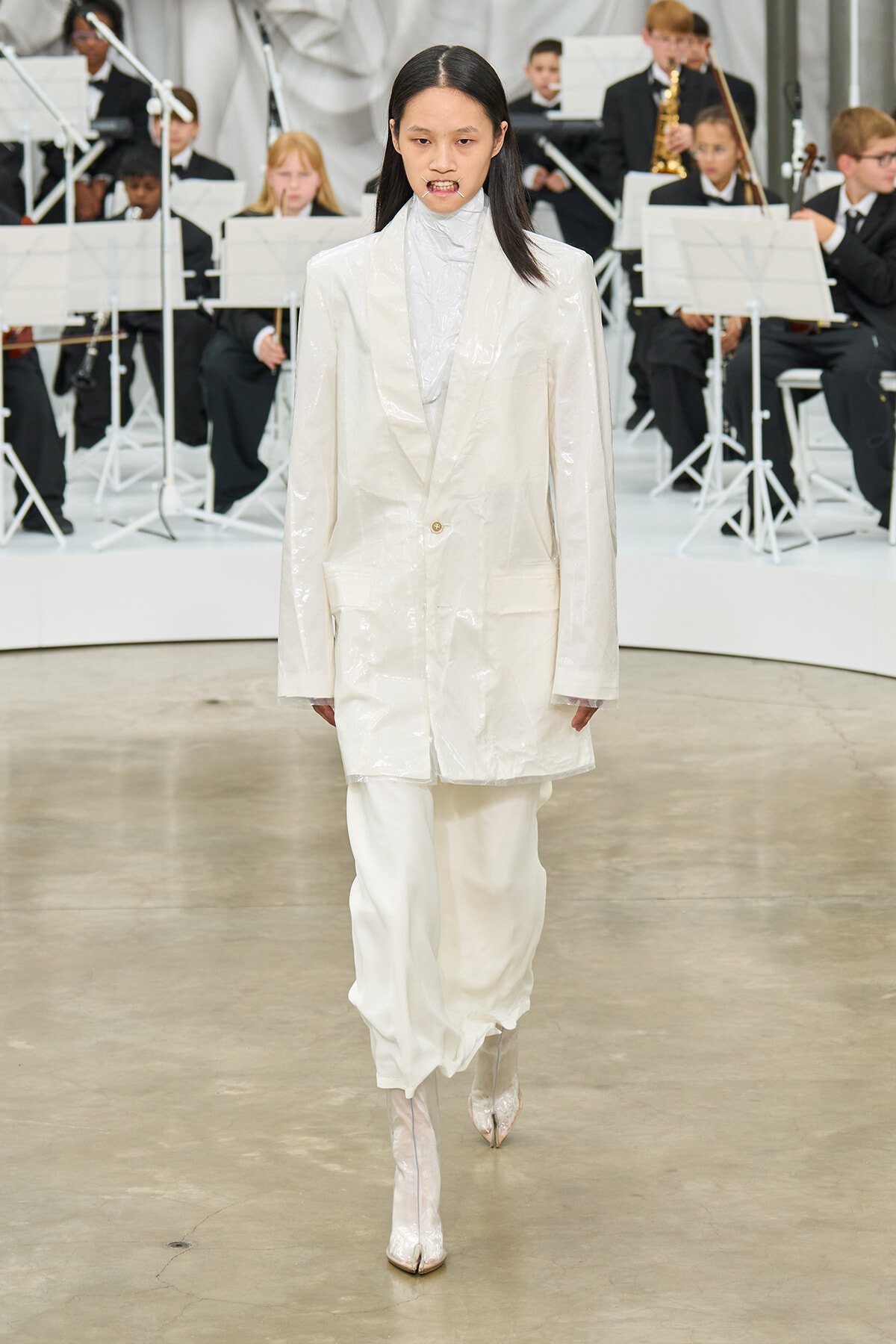 Model in a glossy white suit and matching pants walks a runway, with an orchestra visible in the background.
