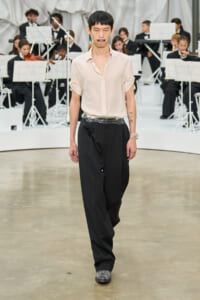 Male model walks a runway in a pale beige shirt and wide black trousers with a metallic belt; an orchestra performs in the background.