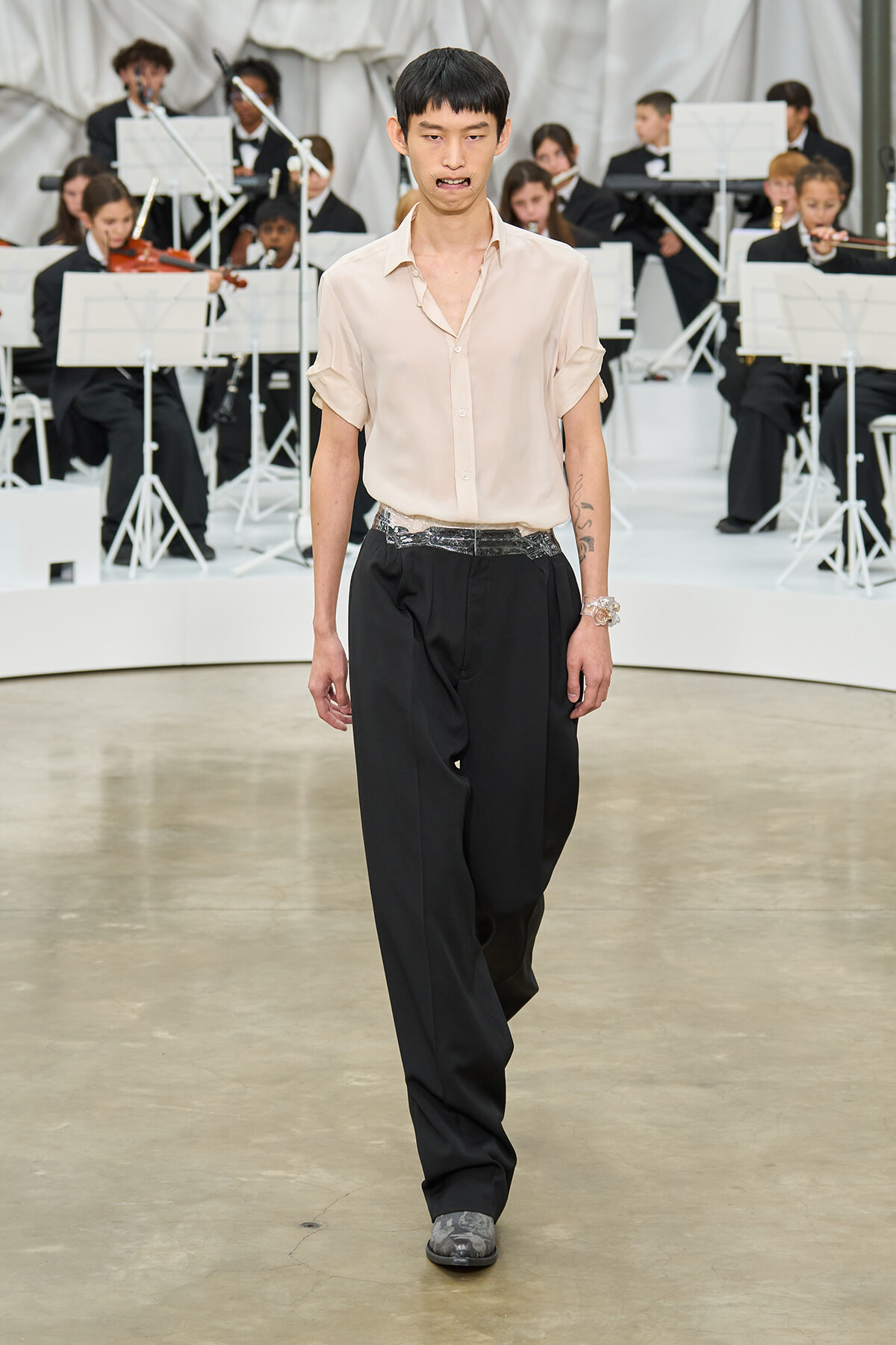 Male model walks a runway in a pale beige shirt and wide black trousers with a metallic belt; an orchestra performs in the background.