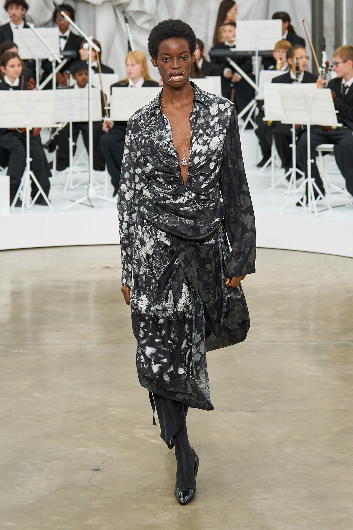 Female model walks a fashion runway in a black-gray floral draped dress with a deep V-neck, wearing a pendant necklace and black boots, as an orchestra audience performs in the background.