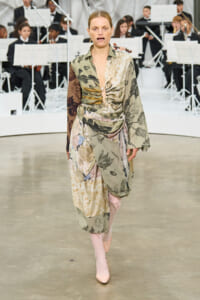 Model walking a fashion runway in a mixed floral print wrap dress with blush pink boots, orchestra visible in the background.