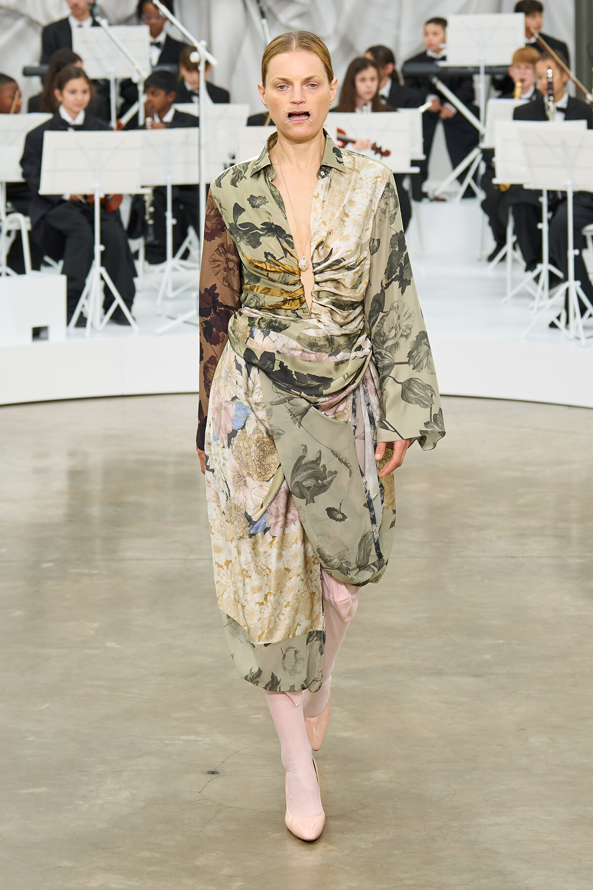 Model walking a fashion runway in a mixed floral print wrap dress with blush pink boots, orchestra visible in the background.