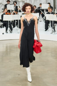 Fashion model walks a runway in a black lace-trimmed jumpsuit, white knee-high boots, and a red handbag; an orchestra plays in the background.