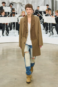 Male model walks a fashion show wearing a tan overcoat, brown scarf, white tee, and patched jeans with tan boots, orchestra in background.