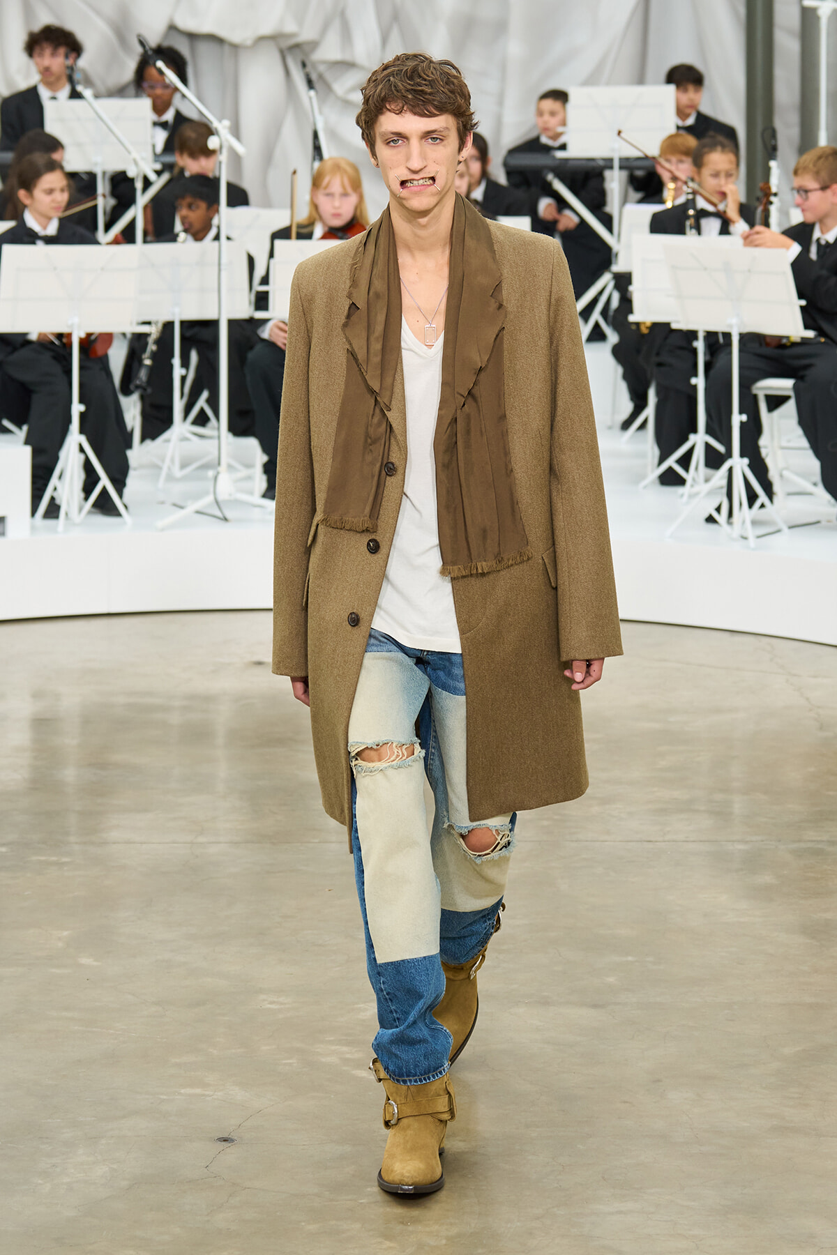 Male model walks a fashion show wearing a tan overcoat, brown scarf, white tee, and patched jeans with tan boots, orchestra in background.