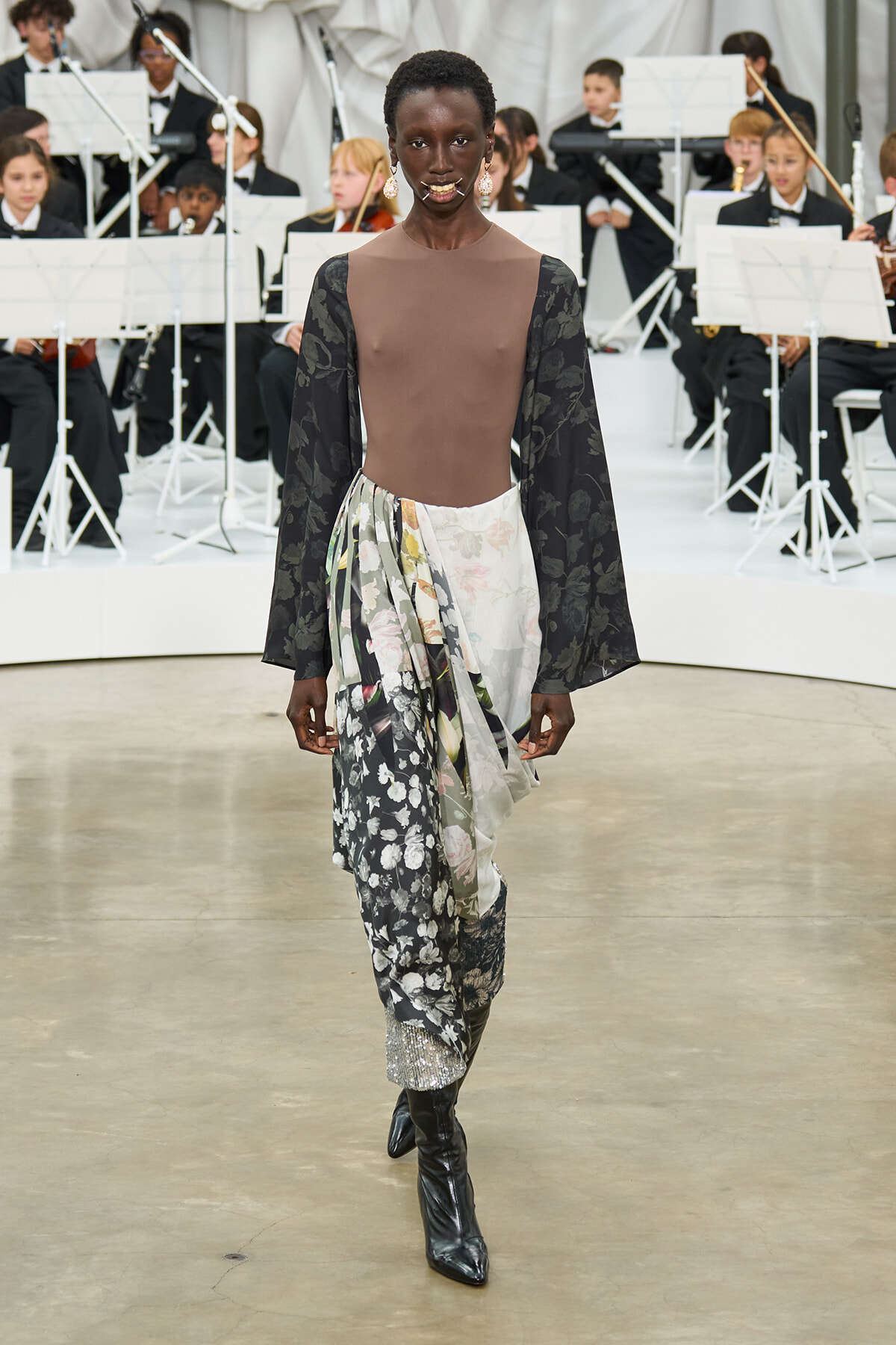 Model walks the runway in a brown sheer bodice with black floral sleeves and a flowing floral wrap skirt, paired with knee-high boots; an orchestra is visible in the background.
