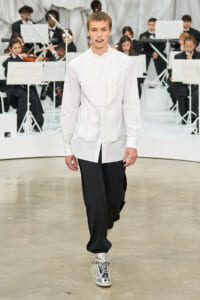 Male model walks a fashion runway in a loose white shirt with a draped bib and black wide-leg pants, wearing metallic silver high-top sneakers as a live orchestra performs in the background.