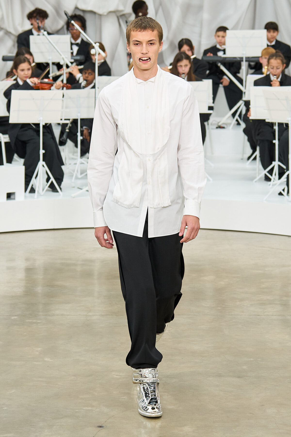 Male model walks a fashion runway in a loose white shirt with a draped bib and black wide-leg pants, wearing metallic silver high-top sneakers as a live orchestra performs in the background.