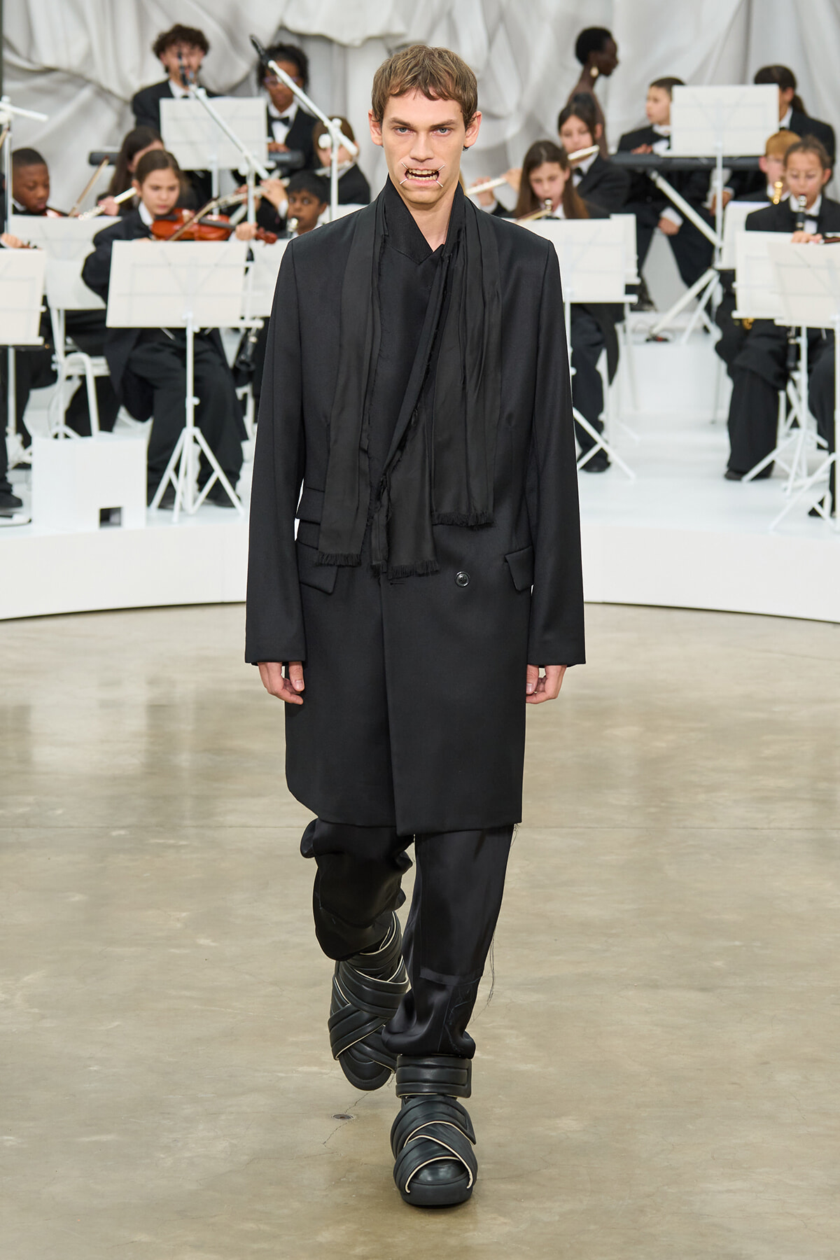 Male model in a black oversized coat and wrapped sandals walks a fashion-show runway, with musicians in black attire performing in the background.