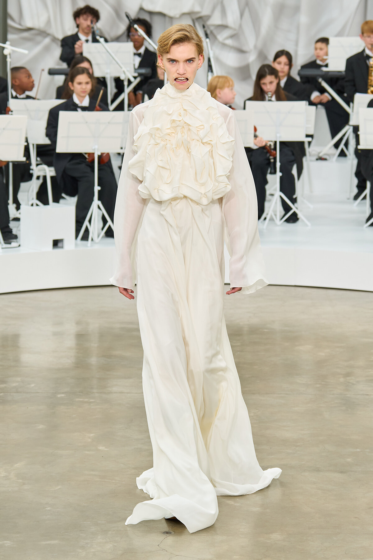 Model in a flowing white gown with a ruffled bodice and sheer sleeves walking a runway, with an orchestra in black ties seated behind.