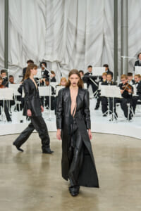 Model walking in a long black leather trench coat on a runway with a seated orchestra in the background.