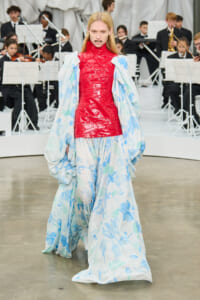 Model walking in a red latex top over a flowing blue-flowered gown on a runway with an orchestra in the background