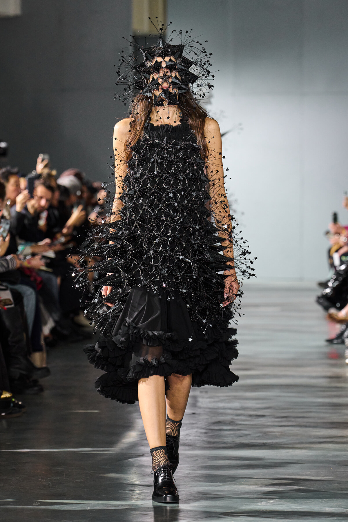 Model walking the runway in a black, ruffled dress densely covered with star-shaped embellishments and a large spiky headpiece. Audience watches from sides.