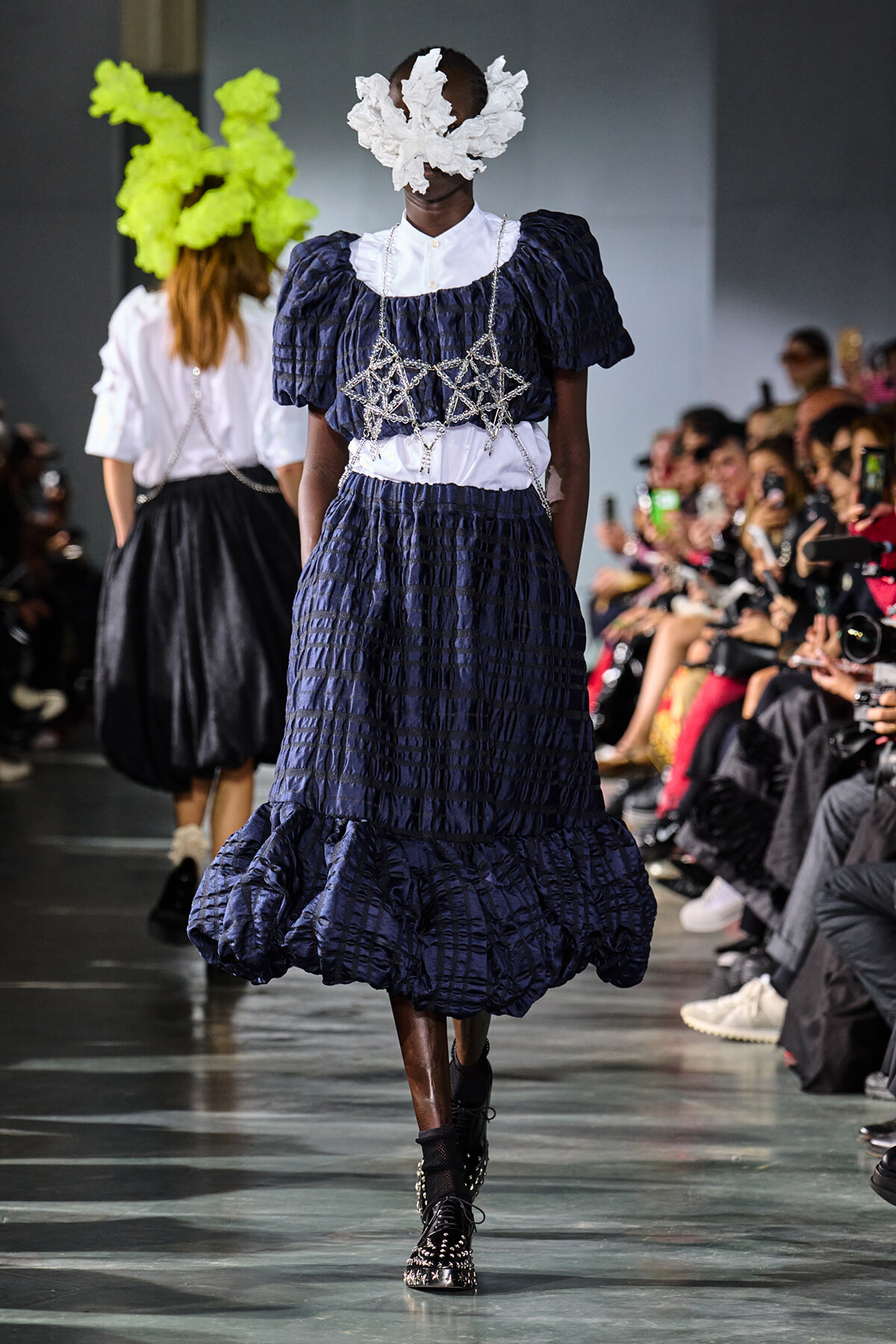 Model walks the runway in a dark blue, puffed gown with a white collar and intricate silver chain harness, paired with a white crumpled headpiece.