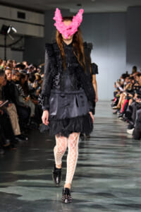 Model walking a runway in a black glittery blazer and satin skirt, with a bright pink feather mask and white lattice tights.