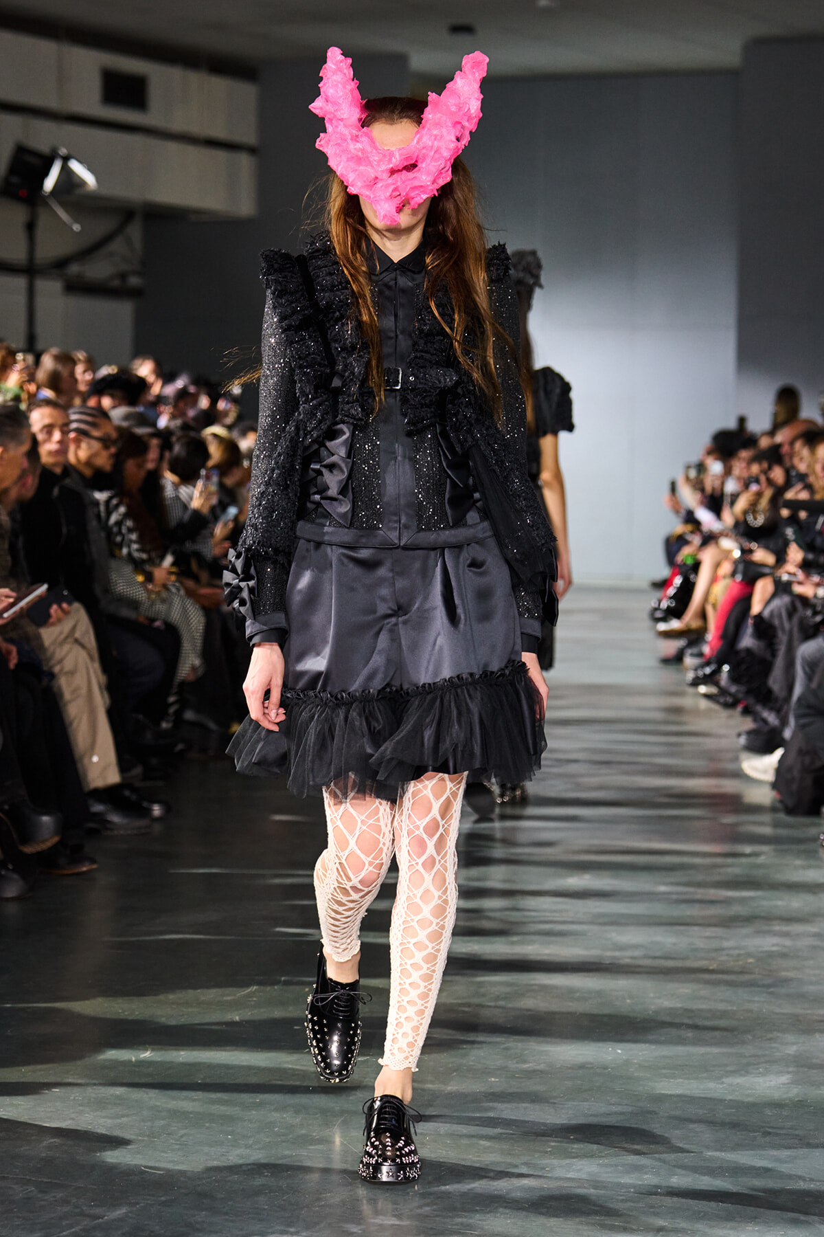 Model walking a runway in a black glittery blazer and satin skirt, with a bright pink feather mask and white lattice tights.