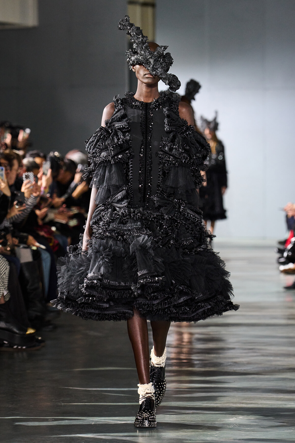 Model on a runway wearing an avant-garde black ruffled gown with a sculptural headpiece; audience visible on both sides.