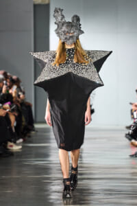 Model walks the runway in a black dress with oversized star-shaped shoulders encrusted with silver studs and a gray sculptural headpiece.