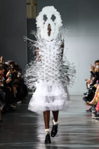 Model walks the runway in a white sculptural gown with silver spiky extensions and a large white mask-like headpiece.