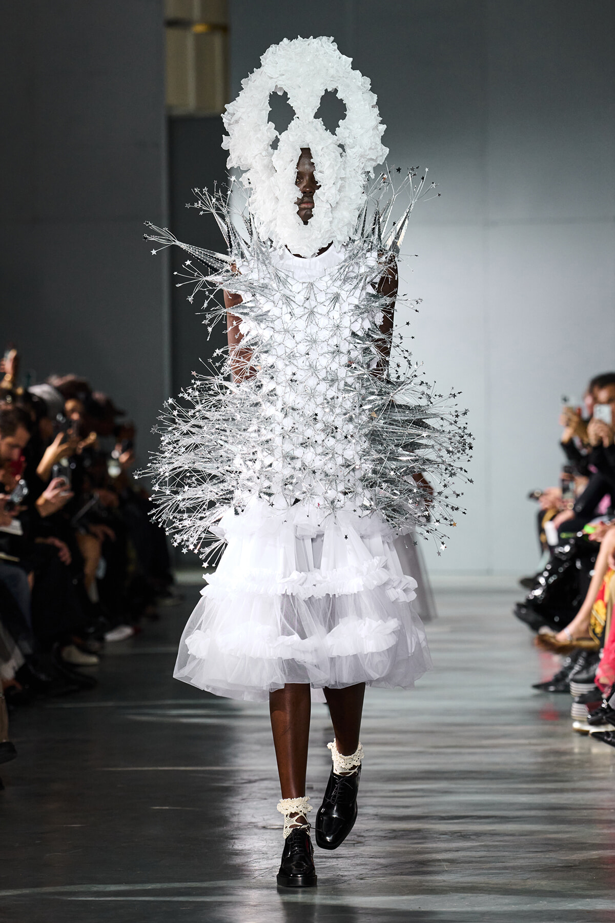 Model walks the runway in a white sculptural gown with silver spiky extensions and a large white mask-like headpiece.